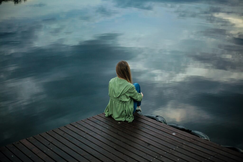 A woman sits on a wooden dock, reflecting by a calm lake under a cloudy sky.