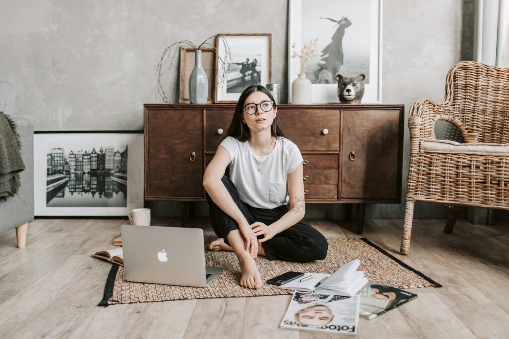 A calm woman with eyeglasses sitting barefoot at home, contemplating and relaxing with a laptop and magazines.