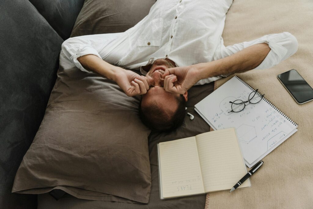 A tired man rubs his eyes while resting on a couch with a notebook, pen, and phone beside him.